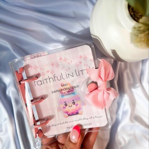 May include: A clear, small binder with pink accents and a pink bow. The binder contains a pink card with the text "Faithful in Little" and a savings challenge graphic of a jar. The text "$450 Savings Challenge" is also visible.