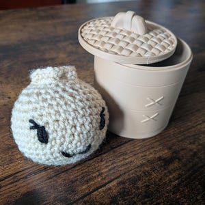 May include: A cream-coloured crocheted dumpling plush toy with black embroidered eyes and a mouth, positioned next to a light beige plastic cylindrical container with a textured lid that mimics a woven basket design.