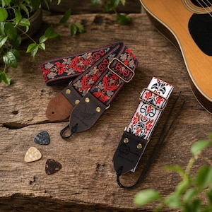 May include: Two guitar straps with floral patterns in red, black, and white, along with three guitar picks. The straps have leather ends and metal hardware. One strap is black with red and gold floral designs, and the other is white with red and black floral designs.