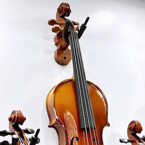 以下が含まれることがあります： 光沢のある木製のバイオリンが壁に掛けられています。バイオリンは黒い弦と茶色のスクロールが付いています。「Phoebe」という名前の木製の壁掛けが見えます。画像は楽器を紹介しています。