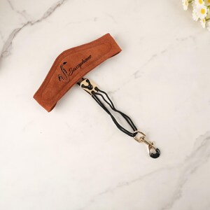 以下が含まれることがあります： 茶色のサックスネックストラップ。パッド入りのネックサポートには「Saxophone」の文字とサックスのグラフィックがプリントされています。黒いコードと金色の金属製の留め金が付いています。