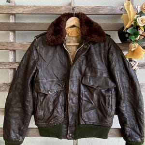 May include: A dark brown leather flight jacket featuring a plush dark brown faux fur collar, a front metal zipper closure, two large flap pockets with snap buttons, and olive green ribbed knit cuffs and a matching ribbed knit waistband.