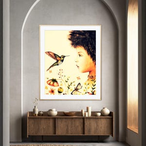 May include: A framed art print showing a watercolour illustration of a person looking at a hummingbird. The artwork includes flowers and butterflies, set against a soft yellow background. The print is displayed in a room with a wooden cabinet and a woven rug.