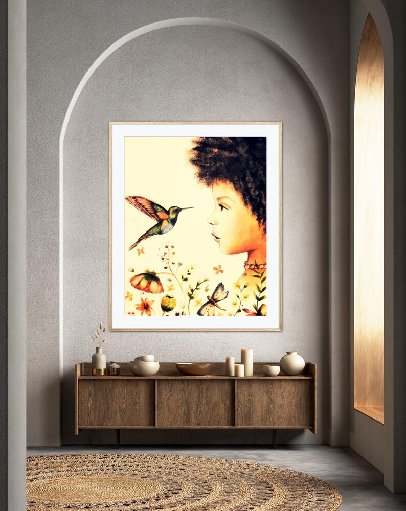 May include: A framed art print showing a watercolour illustration of a person looking at a hummingbird. The artwork includes flowers and butterflies, set against a soft yellow background. The print is displayed in a room with a wooden cabinet and a woven rug.