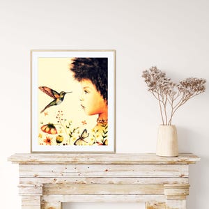 May include: A framed watercolour print depicting a young person with dark curly hair looking at a hummingbird. The artwork includes colourful flowers, butterflies, and a soft yellow background. The print is displayed on a white wall.