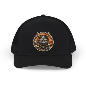 May include: A black trucker hat featuring a circular graphic on the front panel. The design displays a detailed Hannya mask with yellow eyes and horns, set against a background of red sun rays, stylized ocean waves, and a mountain silhouette.