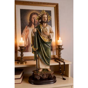 May include: A religious statue of a saint, featuring a green and gold robe, holding a staff. The statue is on a decorative base. A framed picture of Jesus and lit candles are in the background. A rosary and books are also visible.