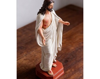 May include: A standing figurine of a bearded man with long brown hair, wearing a draped white robe. The figure has red marks on the chest, hands, and feet, and is mounted on an octagonal reddish-brown base, depicted in a three-quarter view.