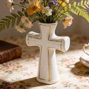 May include: A white ceramic vase in the shape of a cross, with gold trim outlining the cross shape. The vase holds a bouquet of colorful flowers, including orange, yellow, and pink blooms. The vase is a decorative item.