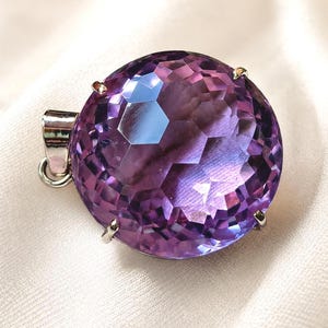 K&ouml;nnte beinhalten: Ein runder, facettierter violetter Amethyst-Anh&auml;nger in einer silberfarbenen Metallfassung mit Krappen. Der Edelstein ist im Brillantschliff mit zahlreichen geometrischen Facetten geschliffen, die das Licht reflektieren, und besitzt eine &Ouml;se.