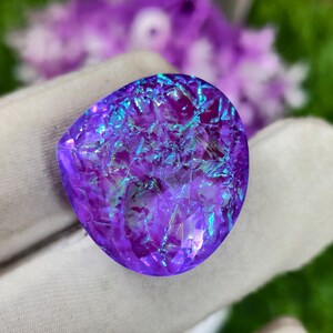 May include: A faceted, iridescent gemstone with a vibrant purple hue, accented by flashes of blue and green. The stone has a unique, irregular shape and a textured surface, reflecting light. The background features blurred purple flowers and green foliage.