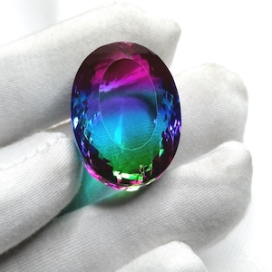 May include: A multi-coloured oval gemstone, displaying hues of purple, blue, and green. The gem is held against a white fabric, highlighting its polished surfaces and internal sparkle. This gemstone is a striking example of fine jewellery.