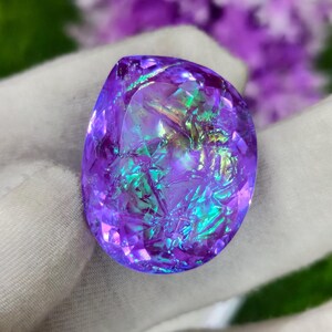 May include: A faceted, oval-shaped gemstone with a vibrant purple hue, displaying iridescent flashes of green and blue. The gem is set against a soft, white fabric, with blurred purple flowers in the background. The gemstone's surface has a fractured, crystalline appearance.