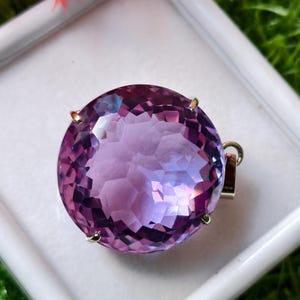 K&ouml;nnte beinhalten: Ein runder, facettierter violetter Edelsteinanh&auml;nger in einer silberfarbenen Metallfassung mit Krappen. Der Stein ist im Brillantschliff gehalten und weist zahlreiche geometrische Facetten auf, die das Licht reflektieren.