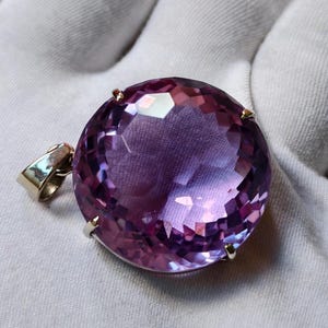 K&ouml;nnte beinhalten: Ein runder, facettierter violetter Edelsteinanh&auml;nger in einer einfachen Metallfassung mit Krappen. Der Stein zeigt einen tiefvioletten Farbton mit Lichtreflexen auf der geschliffenen Oberfl&auml;che. Eine kleine Metall&ouml;se ist seitlich angebracht.