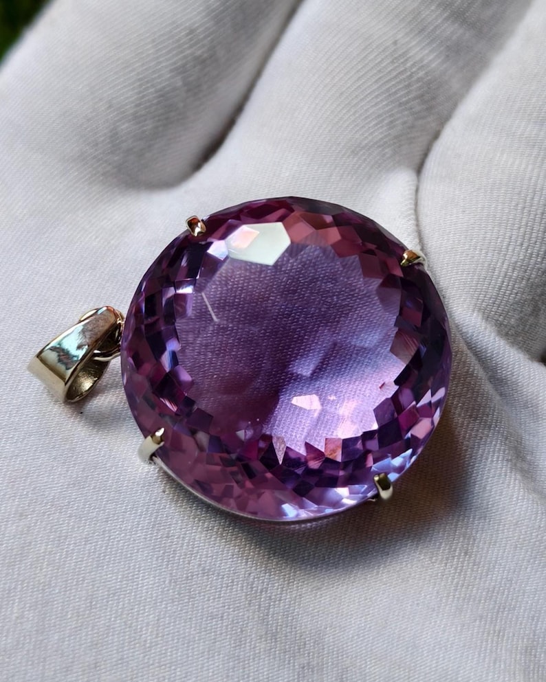 K&ouml;nnte beinhalten: Ein runder, facettierter violetter Edelsteinanh&auml;nger in einer einfachen Metallfassung mit Krappen. Der Stein zeigt einen tiefvioletten Farbton mit Lichtreflexen auf der geschliffenen Oberfl&auml;che. Eine kleine Metall&ouml;se ist seitlich angebracht.
