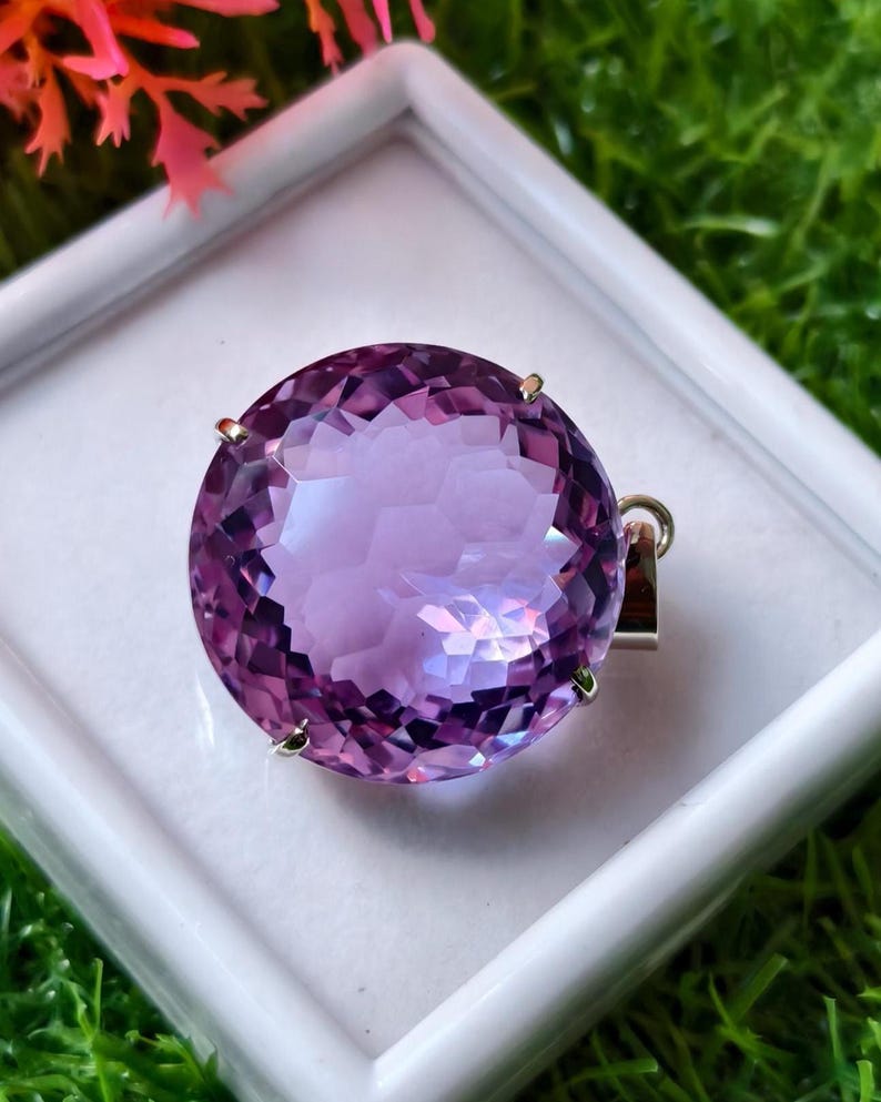 K&ouml;nnte beinhalten: Ein runder, facettierter violetter Amethyst-Anh&auml;nger in einer silberfarbenen Metallfassung mit Krappen. Der Edelstein hat einen Rundschliff mit zahlreichen reflektierenden Facetten, die f&uuml;r ein lichtfangendes Aussehen sorgen. Eine kleine &Ouml;se ist angebracht.