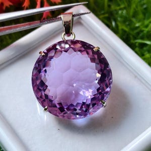 K&ouml;nnte beinhalten: Ein runder, facettierter violetter Amethyst-Anh&auml;nger in einer silberfarbenen Metallfassung mit Krappen. Der Edelstein weist einen komplexen geometrischen Schliff auf, der das Licht reflektiert, und ist an einer silberfarbenen Metall&ouml;se befestigt.