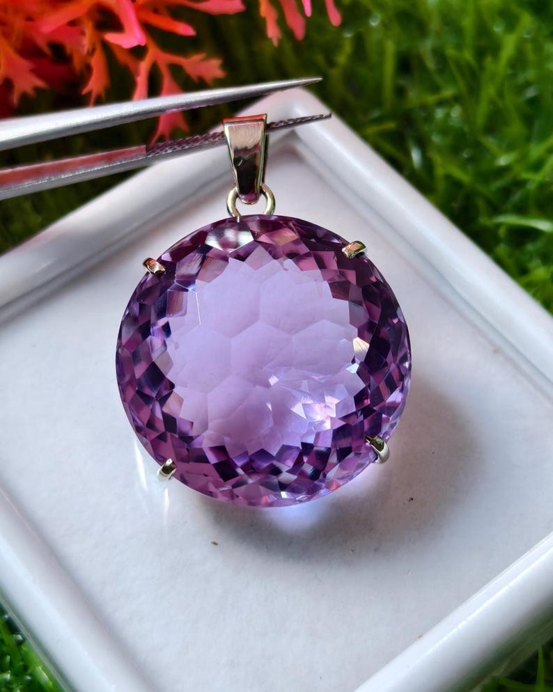 K&ouml;nnte beinhalten: Ein runder, facettierter violetter Amethyst-Anh&auml;nger in einer silberfarbenen Metallfassung mit Krappen. Der Edelstein weist einen komplexen geometrischen Schliff auf, der das Licht reflektiert, und ist an einer silberfarbenen Metall&ouml;se befestigt.