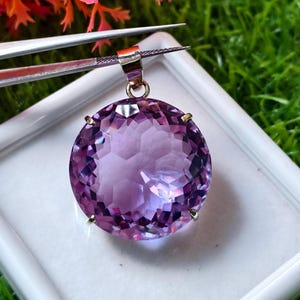 K&ouml;nnte beinhalten: Ein runder, facettierter violetter Amethyst-Anh&auml;nger in einer metallischen Krappenfassung. Der Edelstein weist einen Rundschliff mit zahlreichen reflektierenden Facetten auf, die f&uuml;r ein lichtfangendes Aussehen sorgen. Oben befindet sich eine kleine Metall&ouml;se.