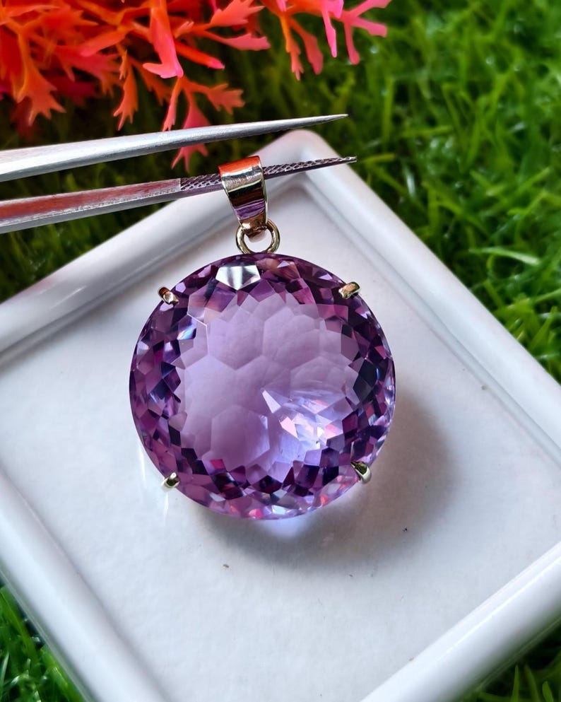 K&ouml;nnte beinhalten: Ein runder, facettierter violetter Amethyst-Anh&auml;nger in einer metallischen Krappenfassung. Der Edelstein weist einen Rundschliff mit zahlreichen reflektierenden Facetten auf, die f&uuml;r ein lichtfangendes Aussehen sorgen. Oben befindet sich eine kleine Metall&ouml;se.