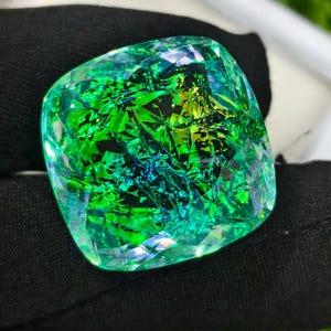 May include: A large, square-cut gemstone with a vibrant, iridescent display of green, blue, and yellow hues. The gem's surface is highly faceted, creating a dazzling play of light and color. The stone is held against a black background.