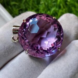 K&ouml;nnte beinhalten: Ein runder, facettierter violetter Edelsteinanh&auml;nger in einer silberfarbenen Metallfassung mit Krappen. Der Edelstein zeigt einen tiefvioletten Farbton mit geometrischen Facetten, die das Licht reflektieren, und besitzt eine Metall&ouml;se.