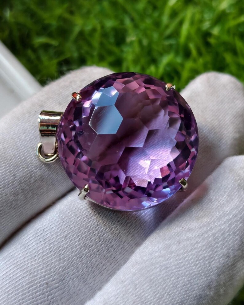 K&ouml;nnte beinhalten: Ein runder, facettierter violetter Edelsteinanh&auml;nger in einer silberfarbenen Metallfassung mit Krappen. Der Edelstein zeigt einen tiefvioletten Farbton mit geometrischen Facetten, die das Licht reflektieren, und besitzt eine Metall&ouml;se.