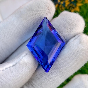 Puede incluir: Una gema facetada de color azul intenso con forma de diamante. La gema se sostiene en una mano enguantada de blanco, con un fondo borroso verde y amarillo. Las facetas de la gema reflejan la luz.