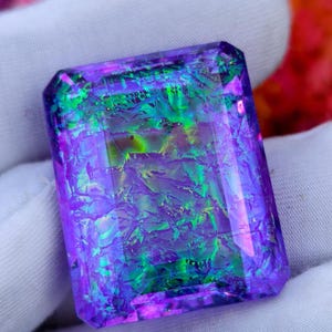 May include: A rectangular gemstone displaying a shimmering array of purple, green, and blue colours. The gem has a textured surface, and is set against a white background. The gemstone is likely a decorative item, possibly for jewellery.