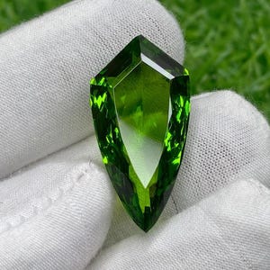 Op de afbeelding: Een gefacetteerde, schildvormige edelsteen met een levendige groene tint. De steen wordt vastgehouden in een witte handschoen, wat de helderheid en glans benadrukt. De achtergrond is een groene, vage weergave.