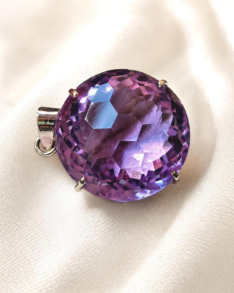 K&ouml;nnte beinhalten: Ein runder, facettierter violetter Amethyst-Anh&auml;nger in einer silberfarbenen Metallfassung mit Krappen. Der Edelstein ist im Brillantschliff mit zahlreichen geometrischen Facetten geschliffen, die das Licht reflektieren, und besitzt eine &Ouml;se.