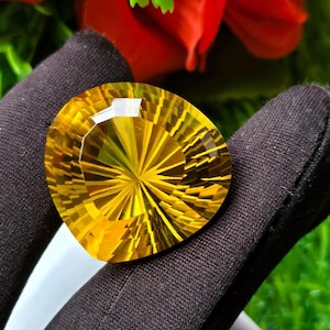 May include: A pear-shaped, faceted yellow gemstone with a radial, sunburst-like pattern cut into its surface. The stone exhibits a bright, golden-yellow hue and is held between two fingers covered in black fabric gloves, showcasing its clarity.