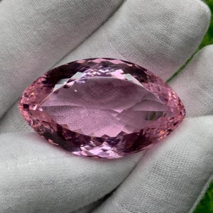 Puede incluir: Una gran gema rosa facetada con talla marquesa. La gema se sostiene en una mano enguantada de blanco, mostrando sus intrincadas facetas y su color rosa claro. La forma de la gema es alargada y simétrica, reflejando la luz.