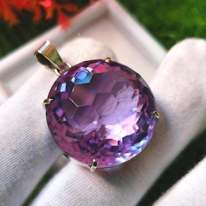 K&ouml;nnte beinhalten: Ein runder, facettierter violetter Edelsteinanh&auml;nger in einer metallischen Krappenfassung. Der Stein zeigt tiefe violette und hellere lavendelfarbene T&ouml;ne mit einer gl&auml;nzenden Oberfl&auml;che. Eine polierte Metall&ouml;se ist an der Oberseite der Fassung befestigt.