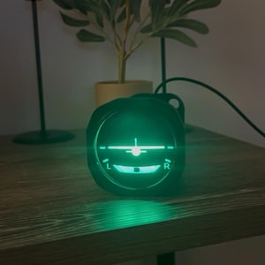 May include: A black aircraft attitude indicator with a glowing green display. The instrument shows a stylized airplane and horizon line. The device is on a wooden surface, with a plant and a decorative metal stand in the background.