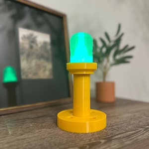 May include: A yellow lamp with a green and blue light. The lamp has a cylindrical base and a round top. The lamp is sitting on a wooden surface. A framed picture and a potted plant are in the background.