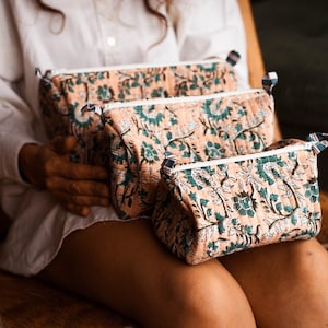 Peut inclure: Trois trousses de maquillage matelassées de différentes tailles. Les sacs présentent un fond pêche avec un motif floral turquoise. Chaque sac a une fermeture éclair blanche et une petite boucle en tissu. Les sacs sont tenus par une personne portant une chemise blanche.