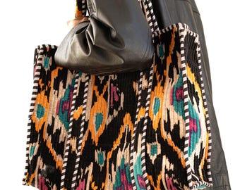 May include: A large tote bag with a colorful, patterned design. The bag features a black background with orange, teal, and pink accents. The straps are black and white striped. The bag is carried over the shoulder.