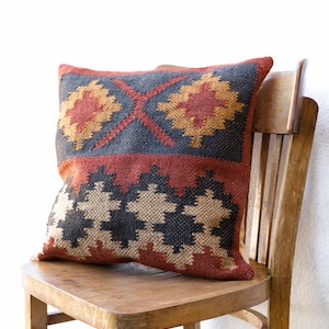 Peut inclure: Un coussin décoratif avec un motif géométrique dans les tons de rouge, noir, beige et or. Le coussin est posé sur une chaise en bois, mettant en valeur sa texture tissée et son design. Les dimensions du coussin ne sont pas visibles.