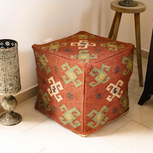 Op de afbeelding: Een vierkante, rode poef met een geometrisch patroon in groen, geel en wit. De poef is gemaakt van een textuurstof en staat op een betegelde vloer. Een decoratieve metalen lantaarn en een kleine houten kruk staan ook in de afbeelding.
