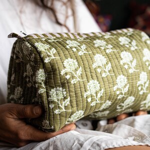 Peut inclure: Trousse de maquillage matelassée vert olive avec un motif floral blanc. Le sac rectangulaire a un accent rayé à une extrémité et une fermeture éclair. Le sac est tenu dans les mains de quelqu'un.