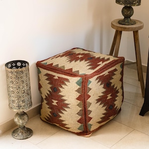 Puede incluir: Un puf cuadrado con un diseño geométrico rojo y beige. También se ven una linterna decorativa de metal y un pequeño taburete de madera. Un mueble de madera oscura está en el fondo.