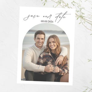 Op de afbeelding: Een witte kaart met een foto van een stel, omlijst door een boog. De kaart leest "Save our date" boven de datum "08/08/2026". Het stel lacht, en de vrouw houdt een mok vast.