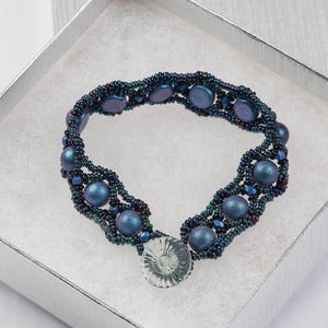 Pode incluir: Pulseira de contas azul escuro e iridescente, apresentada numa caixa de presente prateada. A pulseira apresenta um padrão de contas redondas e planas, mostrando o design e o artesanato.