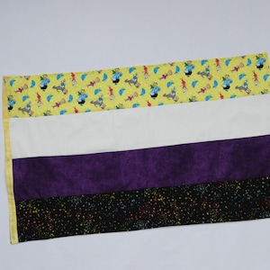 May include: A rectangular quilt with a yellow top border patterned with cartoons, a white stripe, a purple stripe, and a black bottom stripe with colourful speckles. The quilt is edged with yellow fabric.
