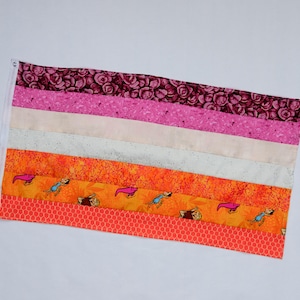 May include: A rectangular fabric flag with horizontal stripes in shades of pink, white, and orange. The top stripe features a floral pattern, while the lower stripes have cartoon dog illustrations. The flag is finished with a white border and grommets.