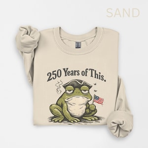 Könnte beinhalten: Sandfarbenes Sweatshirt mit einer Grafik einer grimmigen Froschfigur, die einen Dreispitzhut trägt und eine kleine amerikanische Flagge hält. Der Text "250 Years of This" steht über dem Frosch. Langärmlig.