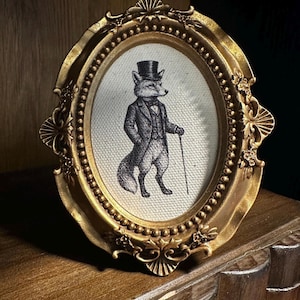 May include: An oval, gold-colored picture frame with a detailed border. Inside, a black and white illustration of a fox in a suit and top hat, holding a cane. The frame is displayed on a wooden surface.