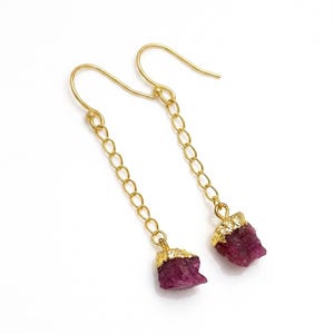 May include: A pair of gold-toned drop earrings featuring a delicate chain link design. Each earring terminates in a raw, deep magenta-coloured stone pendant encased in a textured gold-toned cap, suspended from a simple curved ear wire hook.
